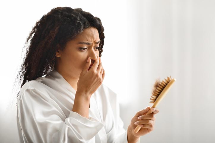 A woman in a white robe holds a brush with hair in it, looking worried and holding her nose with her other hand
